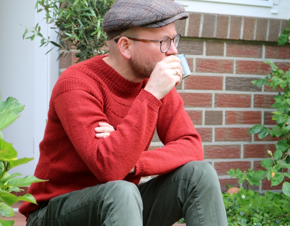 A man in a red handknitted sweater is sitting on the steps outside a door and is taking a sip from a cup.